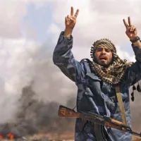 Upornik zoper Gadafijev režim pred gorečim vozilom, ki pripada Gadafijevim silam, kriči “Allahu Akbar!” (“Bog je najbogočnejši!”)