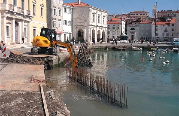 Včeraj se je na ovinku pred palačo Trevisini vdrl zajeten del obale, zato gradbeniki hitijo  čistiti dno in  nameščati betonske opaže (žice iz vode nakazujejo njih rob), do katerih bodo lahko nasuli kamnito  mešanico in tako utrdili obalo