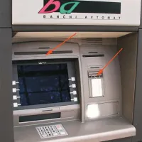 Primer bankomata z nastavljeno napravo; puščica nad monitorjem označuje kamero, ki delno zakriva logotip proizvajalca, druga puščica pa označuje napravo, nameščeno na režo za kartice