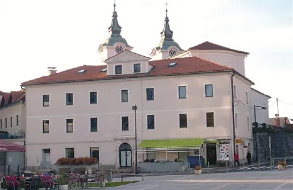 … tik ob njem mogočna meščanska stavba nekdanje Banke Koper, po besedah umetnostnih zgodovinarjev jo v ozadju krasita cerkvena zvonika v slogu mediteransko navdahnjenega baroka, kot prepoznavni simbol občine Postojna.  