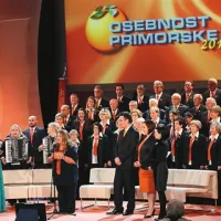 Zborovska dirkgentka Pia Cah pred člani Tržaškega partizanskega pevskega zbora Pinko Tomažič ter med voditeljico prireditve Natašo Benčič in odgovornimi uredniki primorskih medijskih hiš Andrejem Šavkom (Radio Koper), Vesno Humar (Primorske novice ) in