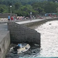 Ob plaži se pozabljen potaplja napihljiv čoln, na njem pa odslužena avtomobilska guma in prazna kanta 