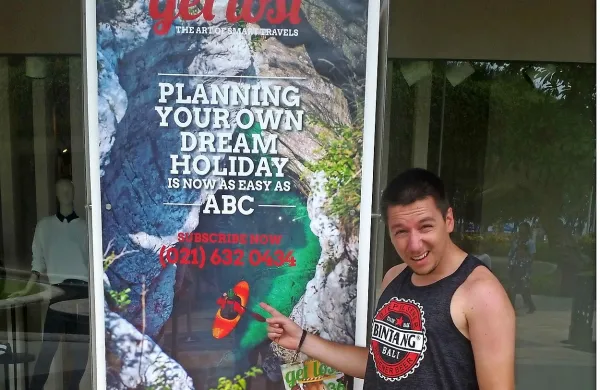 Začudeni David Štulc pred reklamnim plakatom z Velikimi koriti Soče na plaži sanjskega otoka Bali