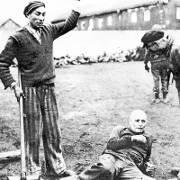  V koncentracijskem taborišču Dachau se osvobojeni taboriščniki norčujejo iz nekdanjega paznika. V ozadju je zid, pred katerim so ameriški vojaki 
postrelili zajete Nemce.