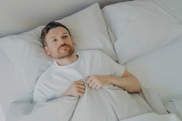 View from above of lonely upset young bearded man lying in bed with open eyes and can't sleep, feeling unhappy and tired, squeezes blanket with hands and being stressed of insomnia. Sleeping problems