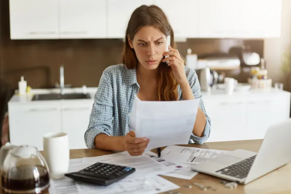 Candid shot of worried young European female dressed casually sitting at kitchen table, holding paper sheet and talking to house and utilities service representative about miscalculation in bills
