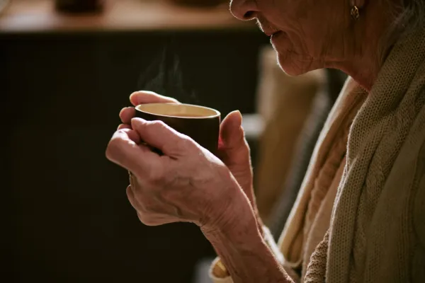 Senior Caucasian woman holding cup with both hands, sipping hot beverage, close up on face and hands, visible wrinkles and textured skin, sitting indoors in relaxed posture
